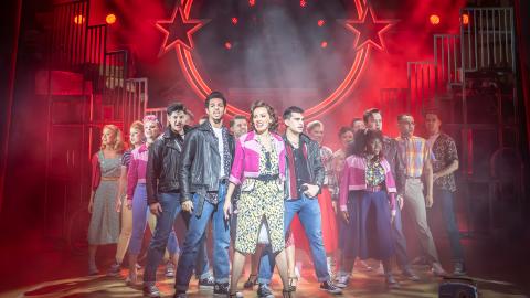 Cast of Grease the Musical on stage with red lights in the background.