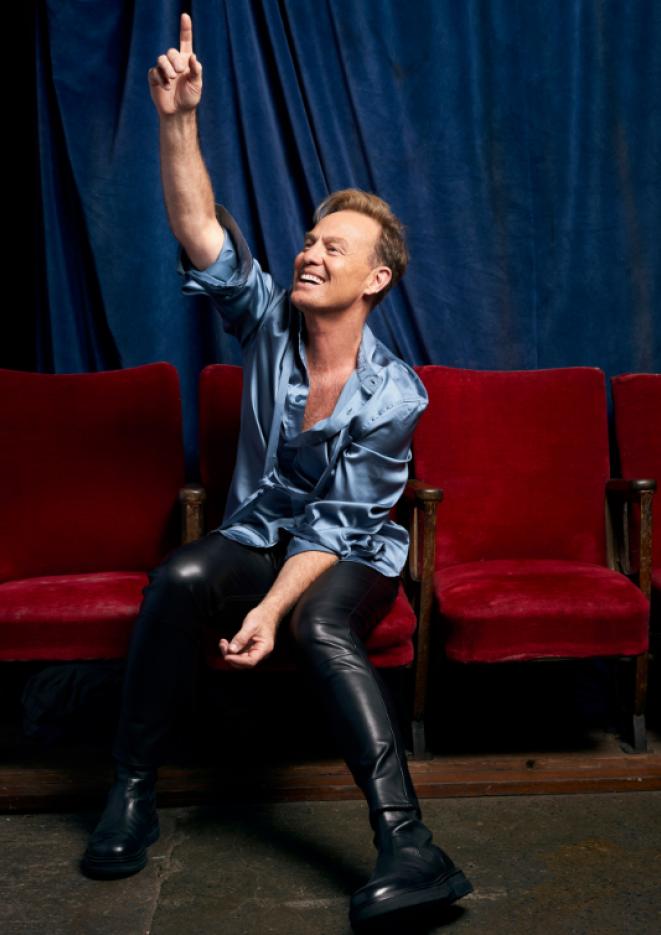 Singer Jason Donovan sits on red theatre seats wearing a blue silk shirt with one arm raised in the air.