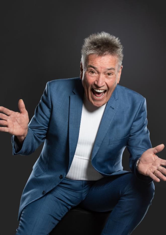 Comedian and actor Billy Pearce sits against a black background wearing a blue suit.