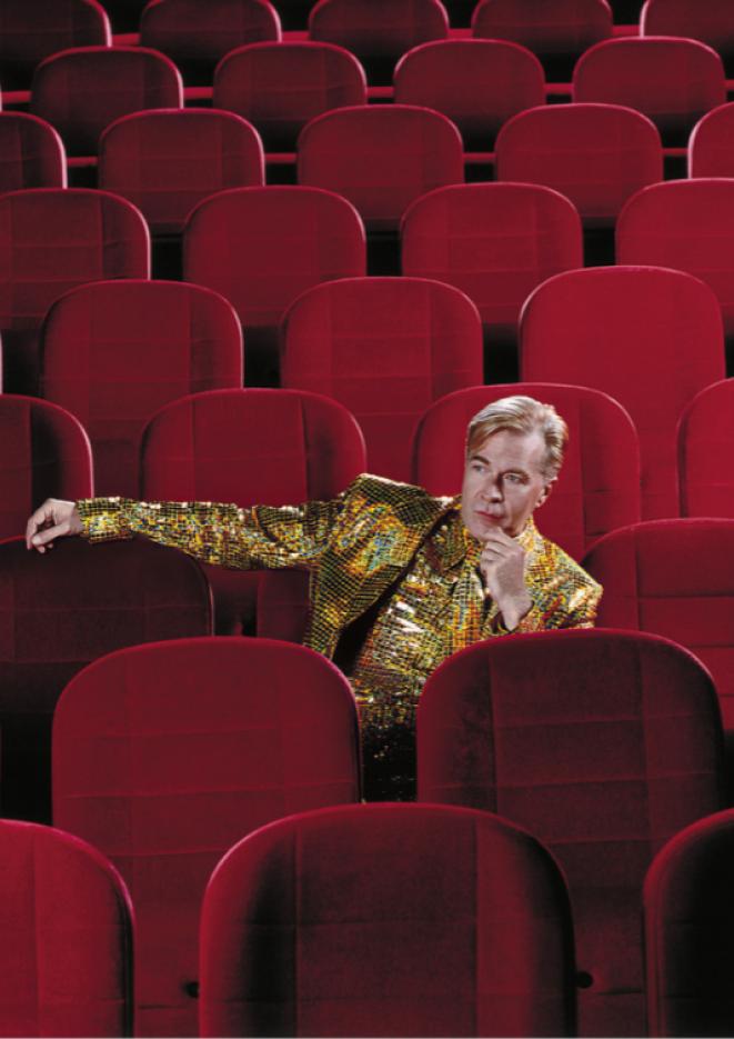 Martin Fry wearing a gold sparkly shirt and jacket, sitting on some red theatre stall seats.