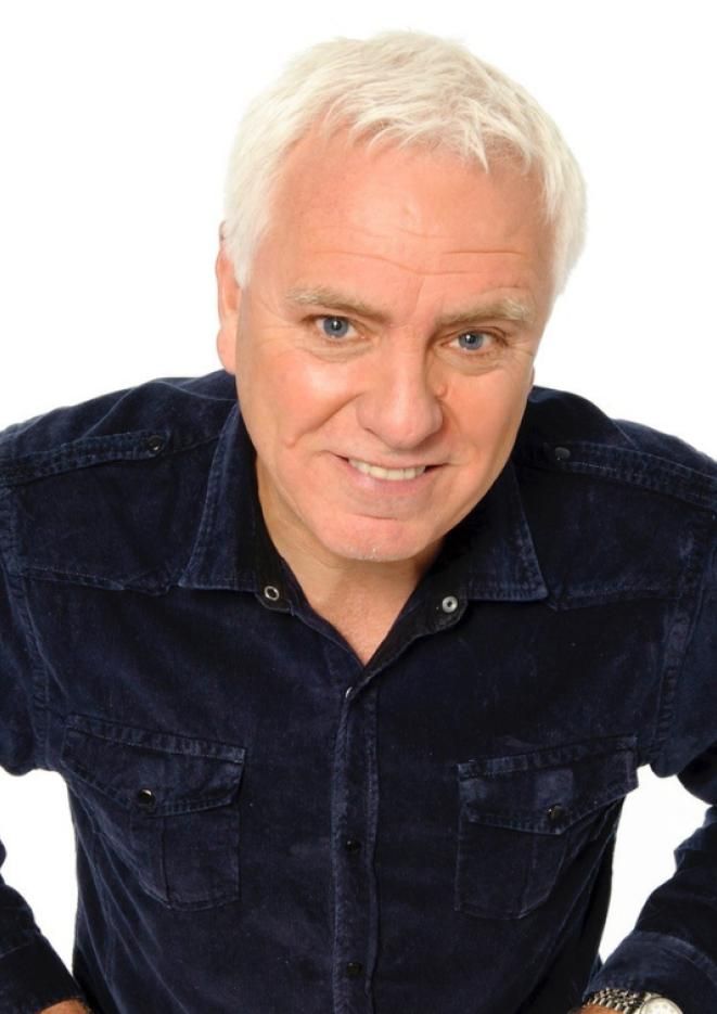 Comedian Dave Spikey stands against a white background looking up at the camera, wearing a dark blue shirt.