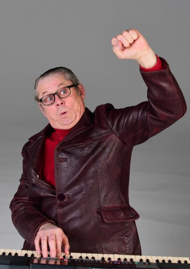 Radio presenter and comedian John Shuttlworth raises his arm in the air while playing a keyboard.