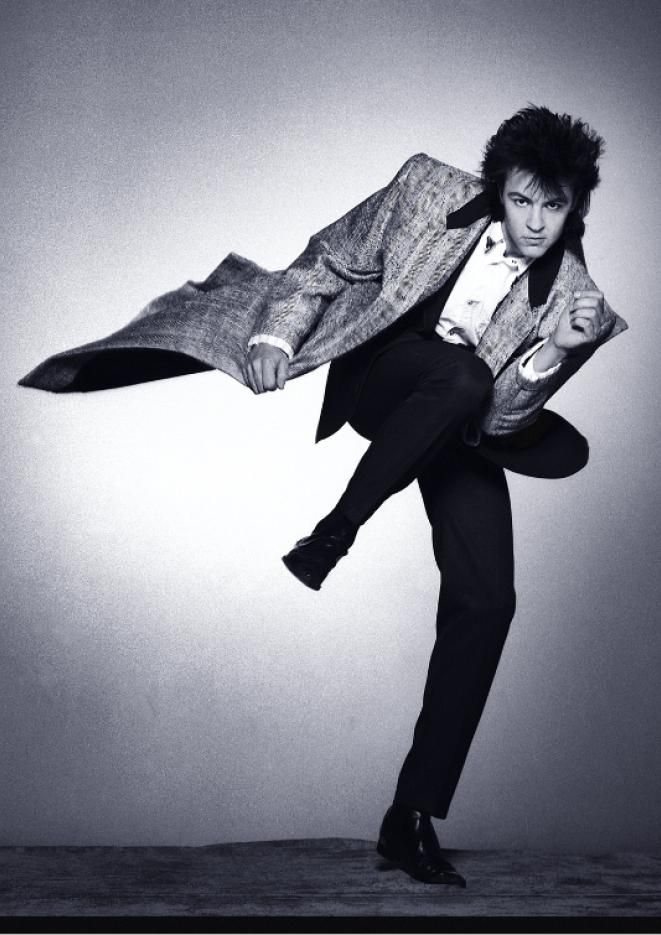 Black and white image of singer Paul Young kicking his leg in the air, wearing a suit and long jacket.