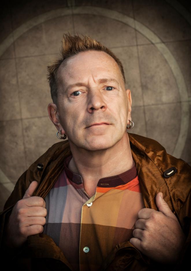 A close up of musician John Lydon wearing a checked shirt and brown leather jacket.