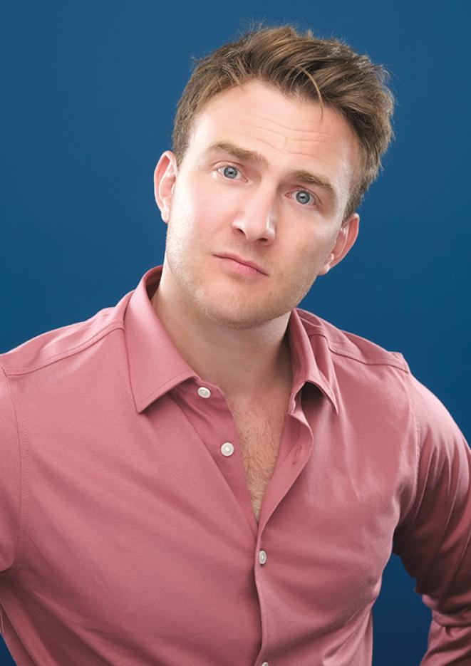 Comedian Tom Houghton poses against a blue background, wearing a salmon coloured shirt and looking directly into the camera.