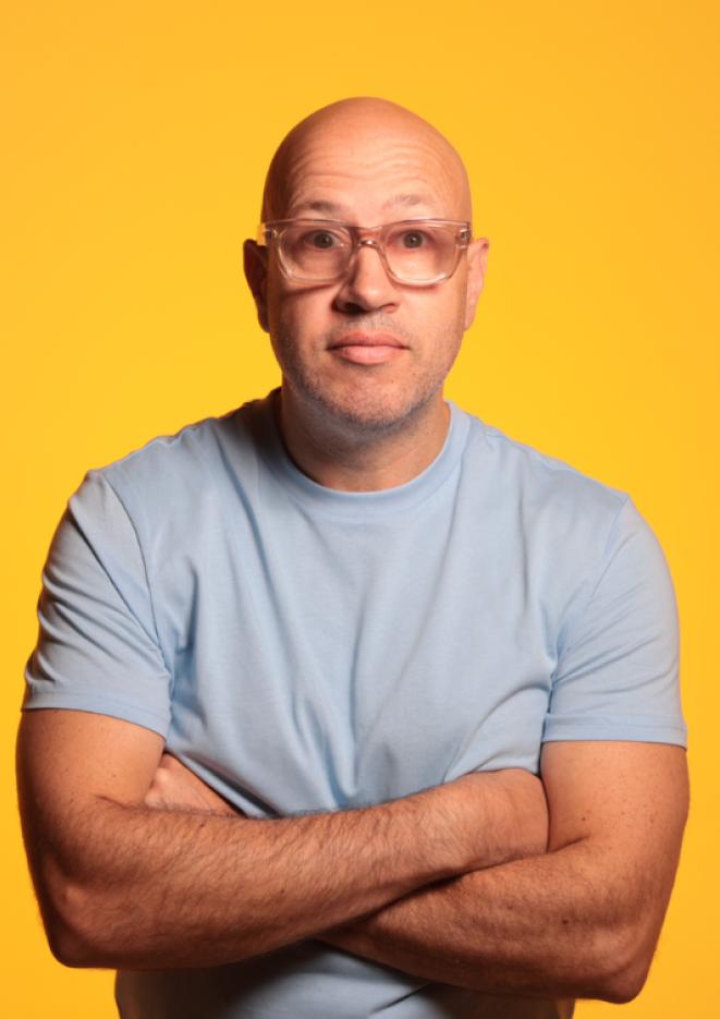 Comedian Sam Avery stands in front of a yellow background with his arms crossed