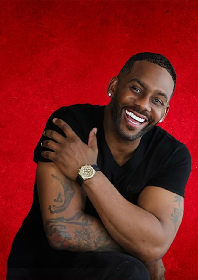 Comedian Richard Blackwood sits in front of a dark red backdrop.