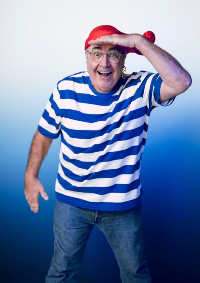 Comedian Danny Baker stands against a faded blue to white background, wearing a striped blue and white shirt and red cap.