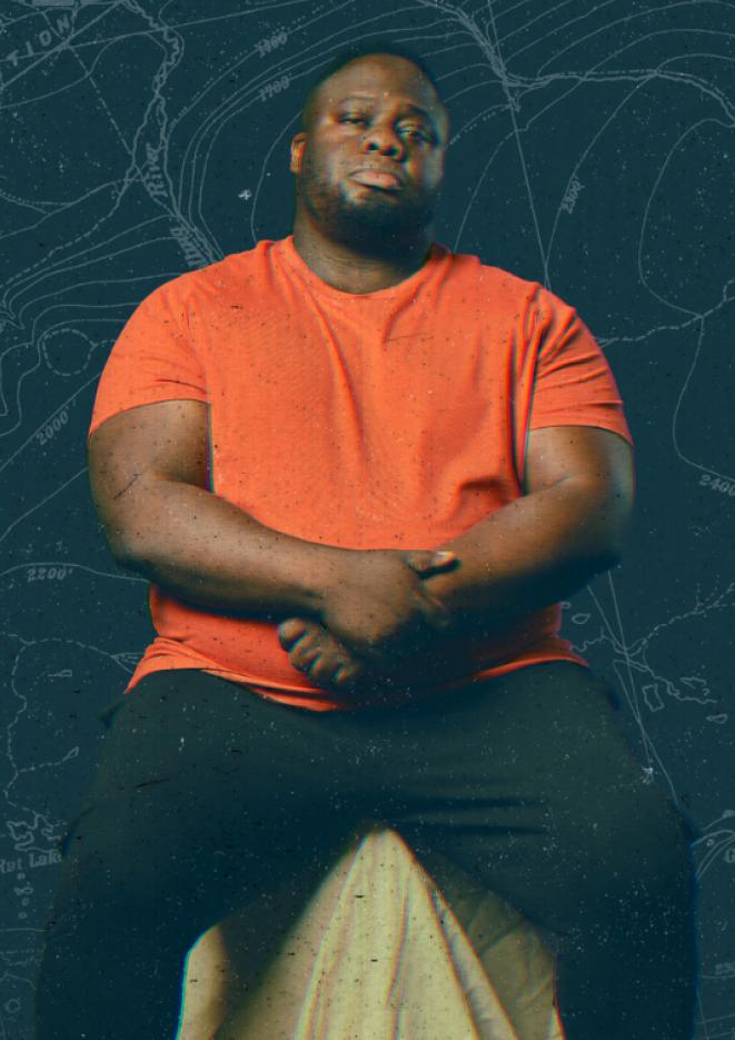 Comedian Nabil Abdulrashid sits in front of a dark blue background wearing an orange tshirt.