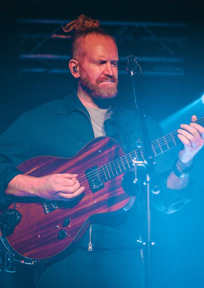 Musician Newton Faulkner plays a red guitar on stage, surrounded by blue stage lighting.