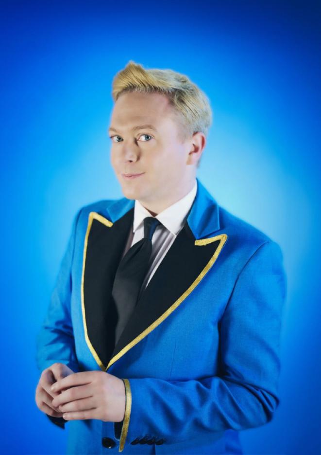 Comedian Stephen Bailey stands against a plain blue background wearing a blue and gold jacket.