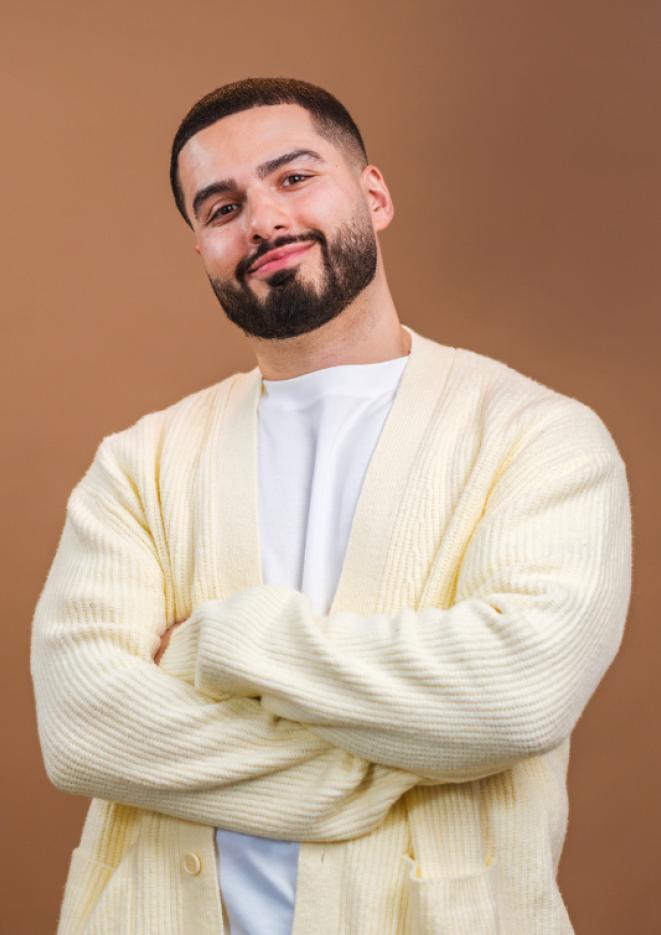 Comedian Kae Kurd stands against a brown background wearing a yellow cardigan and white shirt, with his arms crossed.