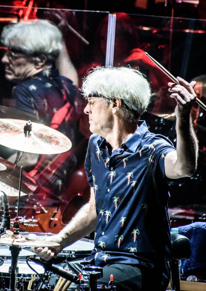 Drummer Stewart Copeland is playing the drums in a blue shirt with red stage lights around him.