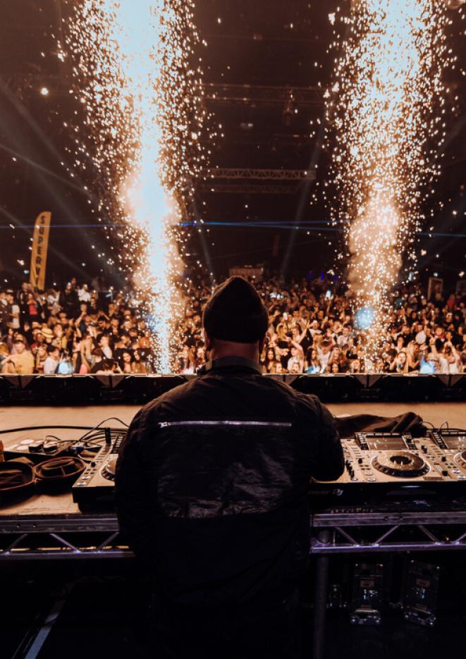 A DJ seen from behind wearing all black in front of decks, a crowd can be seen in the background along with large sparklers.