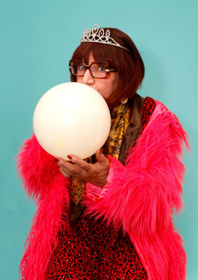 Comedian Barbara Nice sits in front of a blue backdrop wearing a feathered pink jacket while blowing up a balloon.