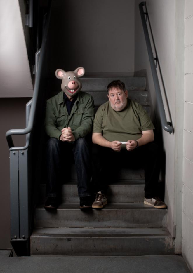 Johnny Vegas and a man in a mouse mask sit on some dimly lit stairs.